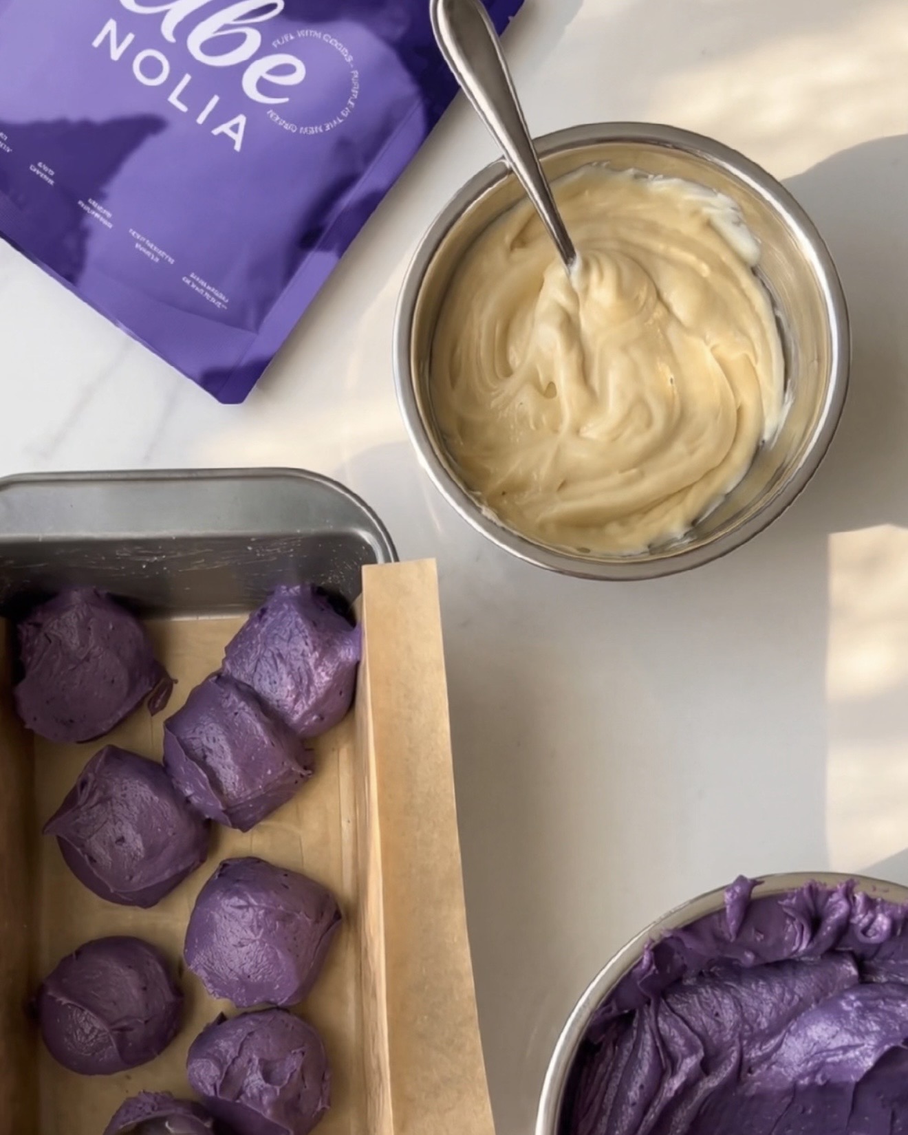 Finished homemade ube halaya, vanilla batter and scoops of purple ube batter in a loaf pan next to a pouch of Ube Nolia ube powder