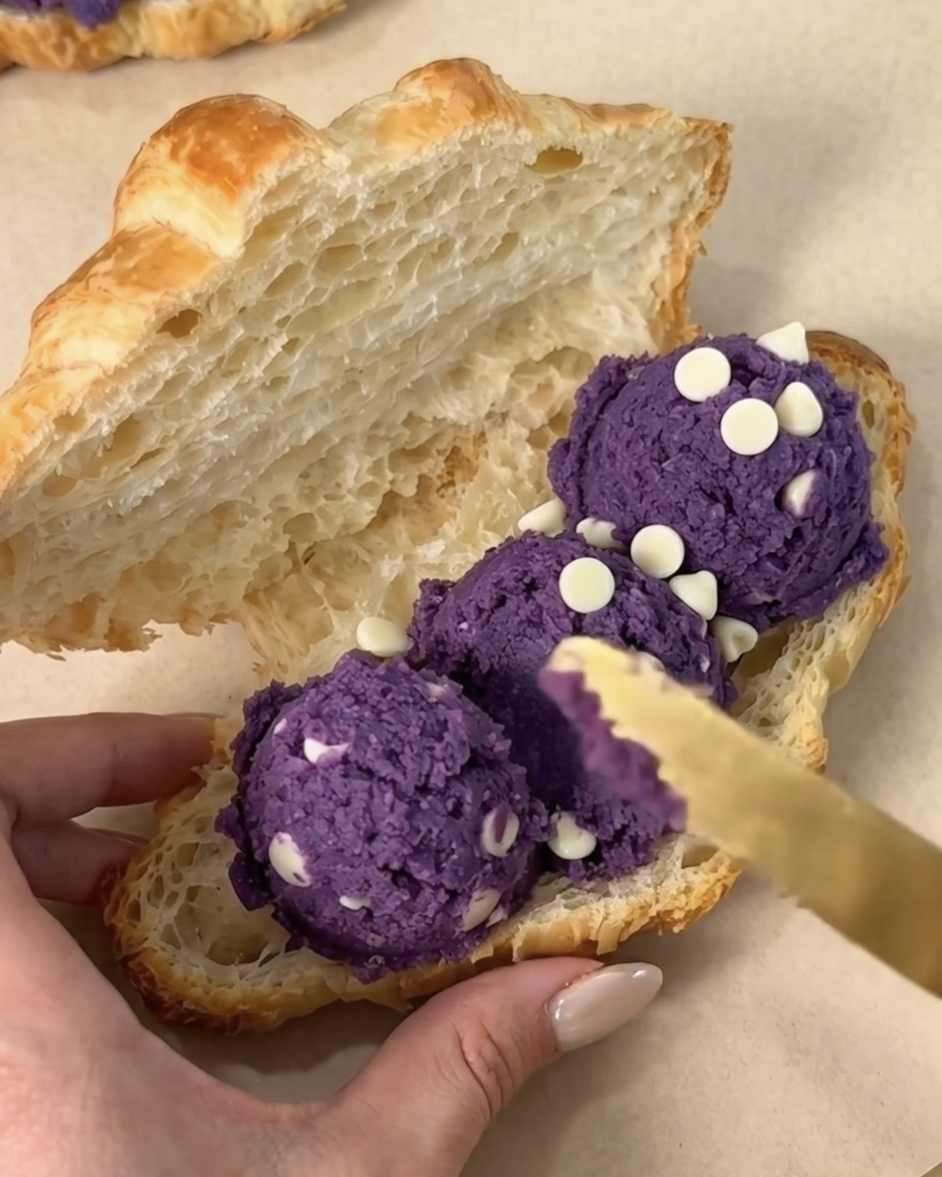 Espalhando colheres de massa de biscoito de ube roxo (inhame roxo) com pepitas de chocolate branco UBE NOLIA® dentro de um croissant de manteiga de padaria fatiado para fazer um ube crookie