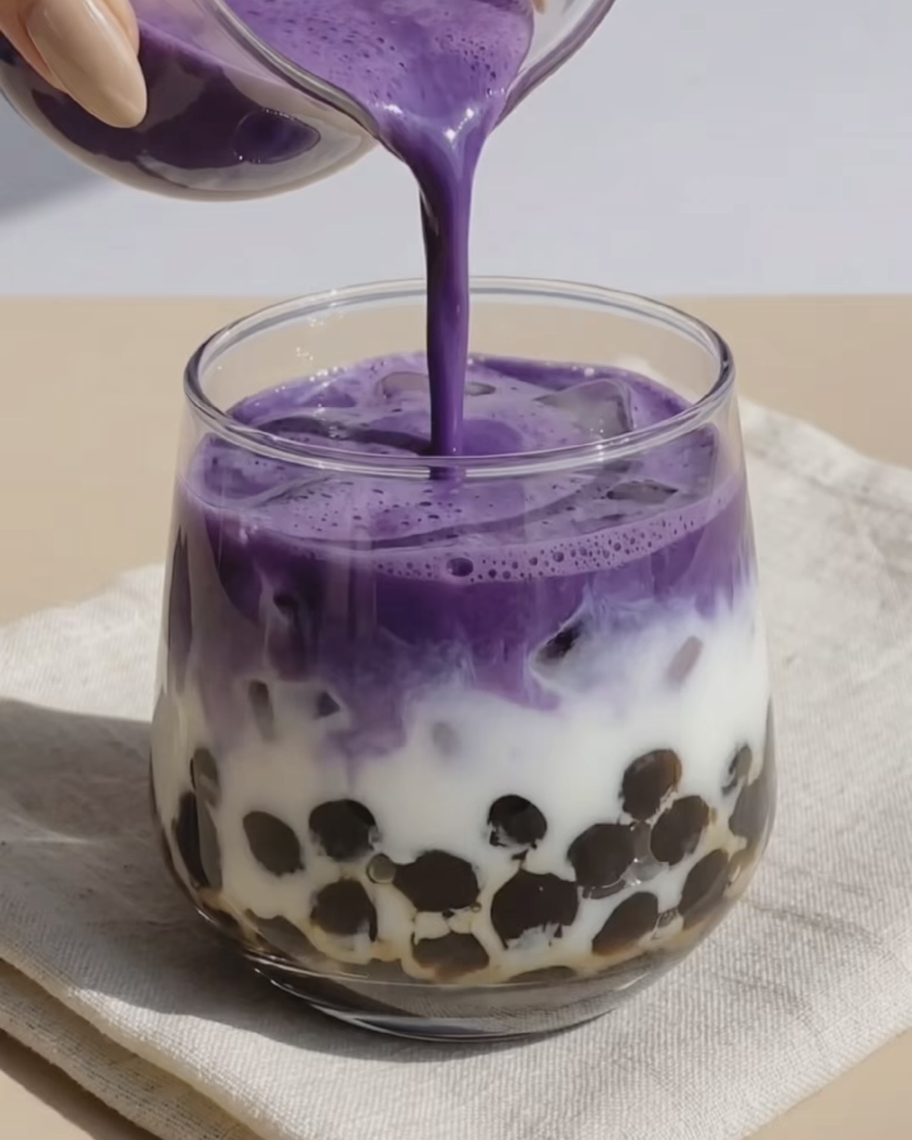 Purple ube oat milk being poured from a small jug into a glass of black tapioca pearls and brown sugar syrup by Ube Nolia