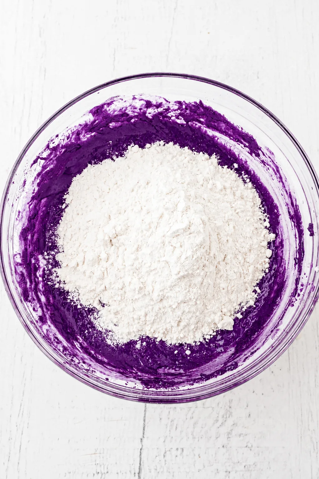 Flour added on top of the deep purple ube white chocolate brownie batter in a glass bowl, ready to fold with Ube Nolia powder