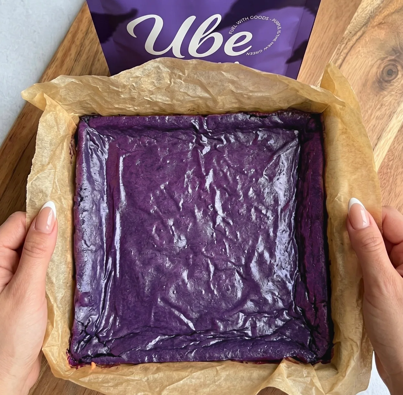Deep purple ube brownie batter layered in a parchment-lined square pan next to an Ube Nolia powder pouch